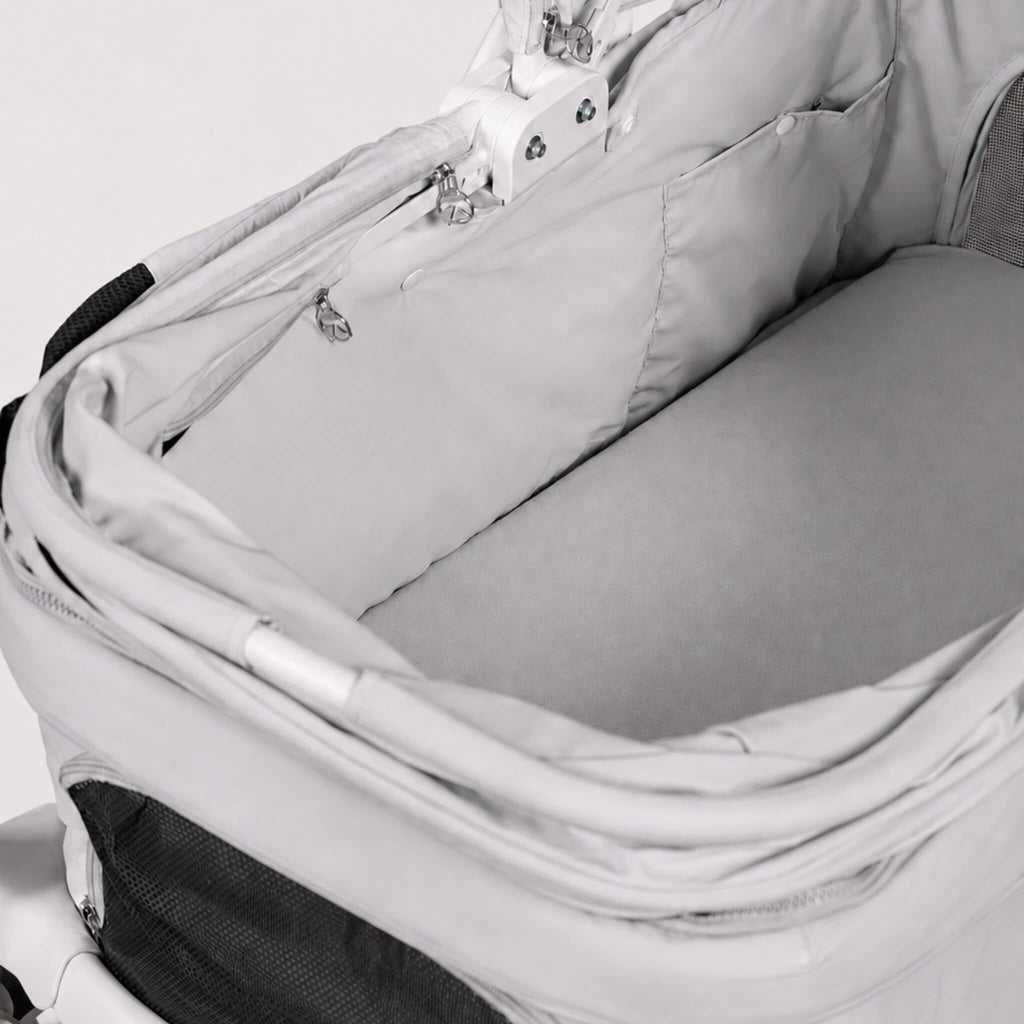 Close-up of a light gray stroller bassinet insert with a padded bottom base and zippered pocket, shown inside a stroller frame.