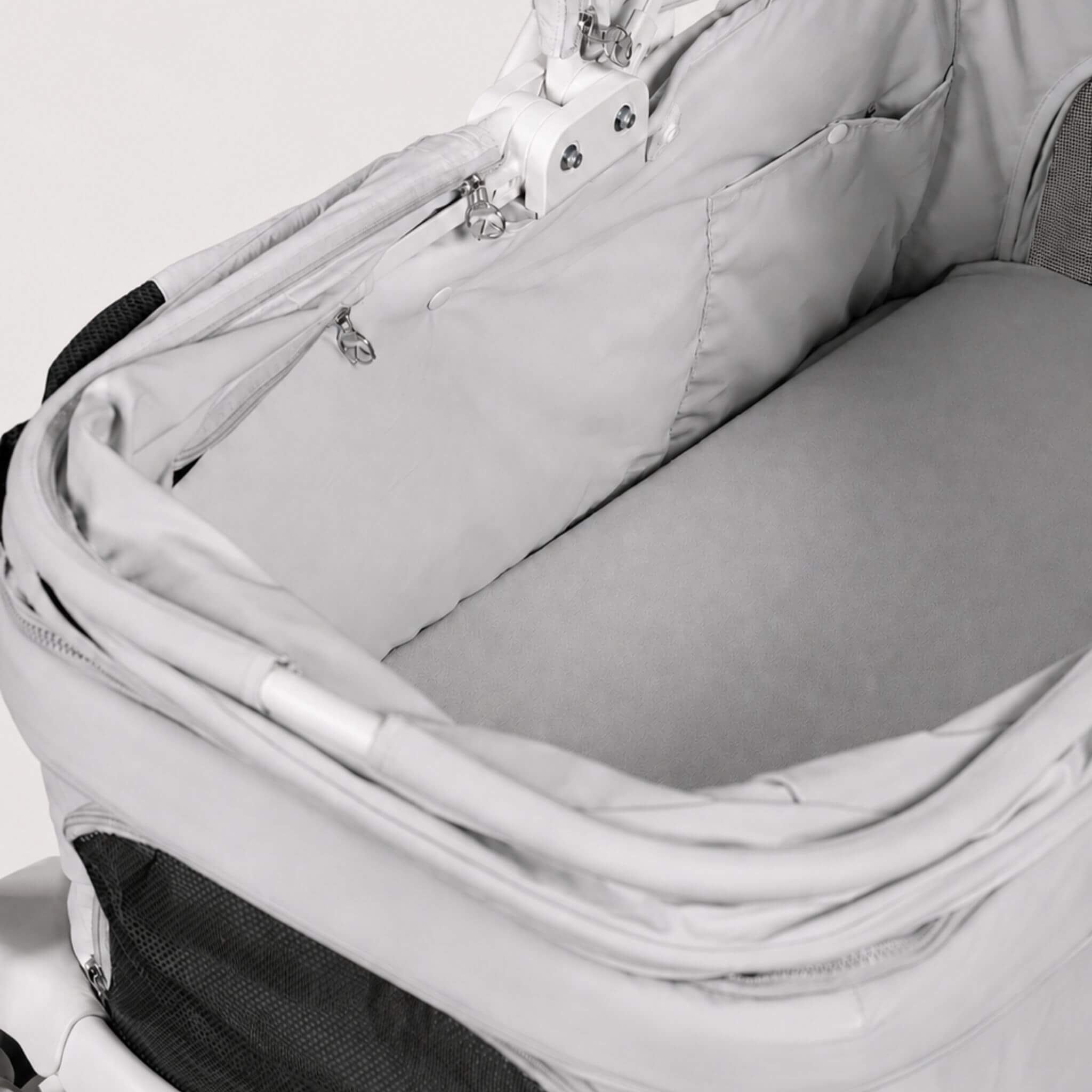 Close-up of a light gray stroller bassinet insert with a padded bottom base and zippered pocket, shown inside a stroller frame.